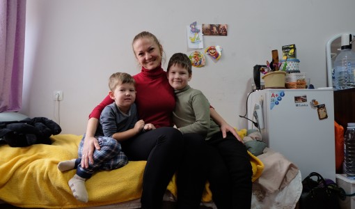 En kvinna och hennes två barn sitter på en säng. De tittar in i kameran och ler.