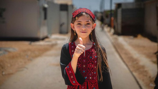 En flicka står utomhus i ett flyktingläger och håller upp tummen.