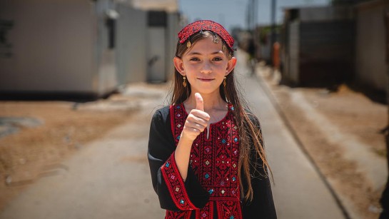 En flicka står utomhus i ett flyktingläger och håller upp tummen.
