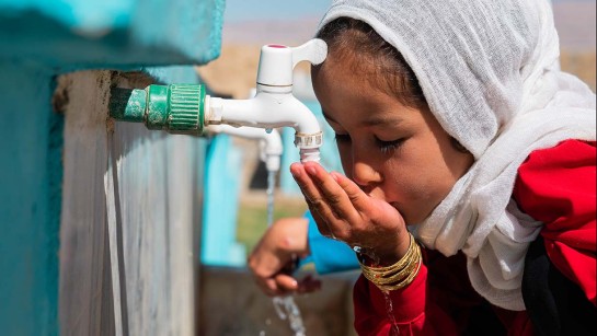 En liten flicka dricker rent vatten från en kran utomhus.
